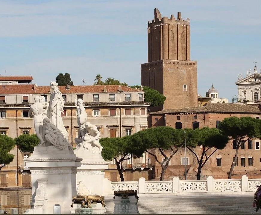 An editorial style image depicting a historic cityscape, featuring prominent white marble statues in the foreground against a backdrop of older brick buildings and a tall, ancient tower under a clear sky.