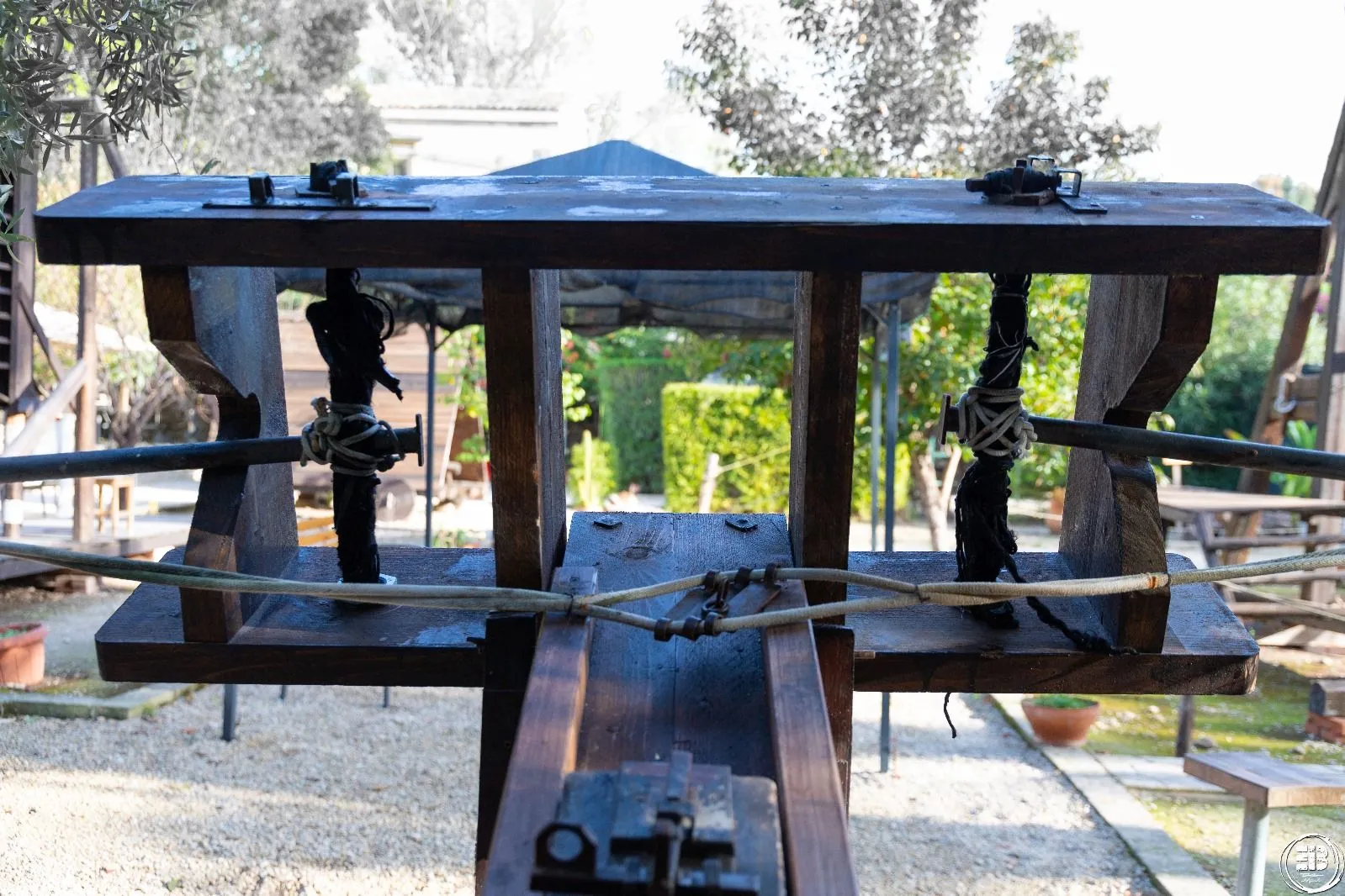 A close-up view of an intricate wooden mechanism, possibly a part of an old machine or exhibit, with ropes and metal fittings. The structure frames a soft-focused background of a lush green garden with hedges and trees under natural daylight.