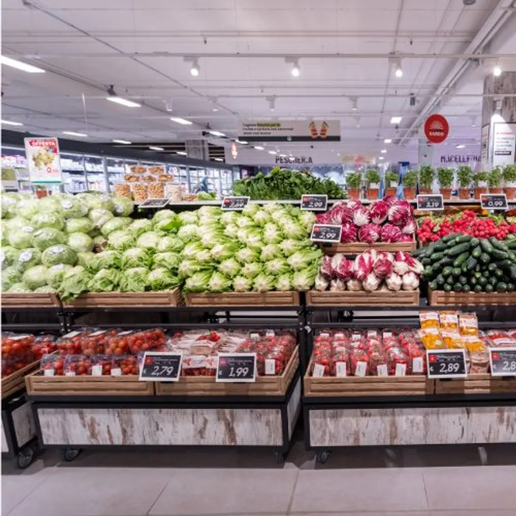 The image is well-composed, providing a clear and comprehensive view of a supermarket's produce section. It benefits from excellent lighting, which highlights the vibrant and natural colors of the fresh produce, contributing to its strong photo-realistic quality. Key elements like the fruits and vegetables in the foreground and mid-ground are sharp and in focus. While standard price tags and background store signage are present, they are integral to an editorial depiction of a retail environment and do not detract from the overall quality or function as primary promotional graphics.