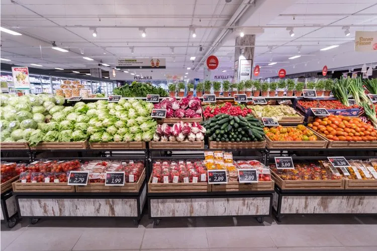 The image is well-composed, providing a clear and comprehensive view of a supermarket's produce section. It benefits from excellent lighting, which highlights the vibrant and natural colors of the fresh produce, contributing to its strong photo-realistic quality. Key elements like the fruits and vegetables in the foreground and mid-ground are sharp and in focus. While standard price tags and background store signage are present, they are integral to an editorial depiction of a retail environment and do not detract from the overall quality or function as primary promotional graphics.