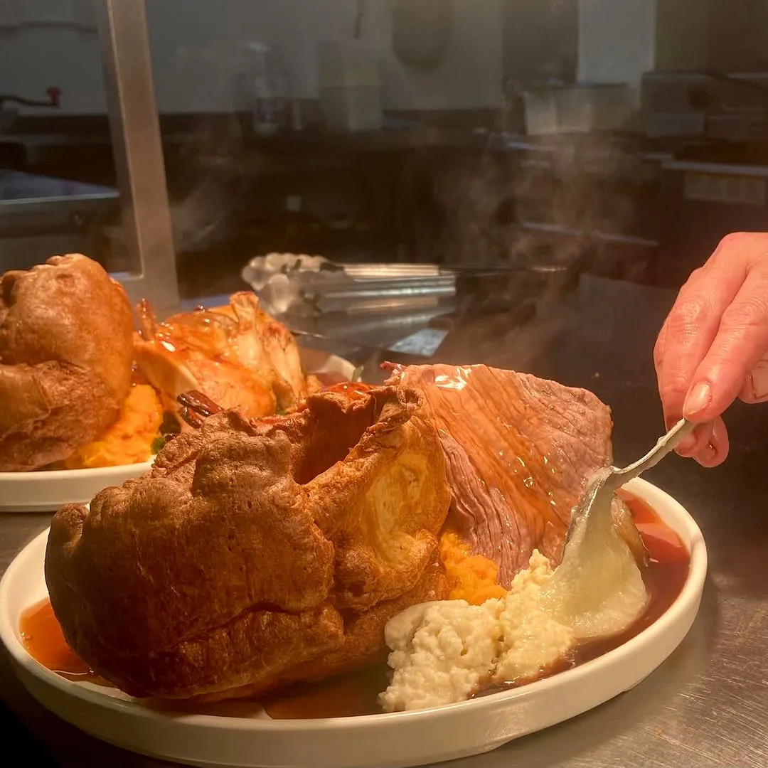 A close-up shot of a steaming hot roast dinner being served. The main elements of the dish, including a large Yorkshire pudding, sliced meat, and mashed potatoes, are clearly visible and in focus. The steam rising from the food adds to its appeal, conveying freshness and warmth. A hand is present, using a spoon to add or adjust an element of the dish, which enhances the dynamic nature of the shot. The colors appear natural, and the image is photo-realistic.