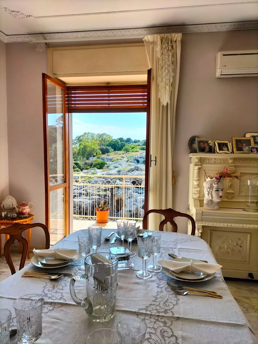 The image is a well-composed editorial-style shot of a dining room with a scenic balcony view. Its strengths include its photo-realistic quality, sharp focus on the main elements like the set table and the landscape, and effective framing that provides depth. However, the lighting presents a challenge due to the high contrast between the bright exterior and the dimmer interior, leading to some underexposure inside and minor overexposure in the sky. The composition could be marginally improved with more balanced framing and careful attention to elements at the edges.