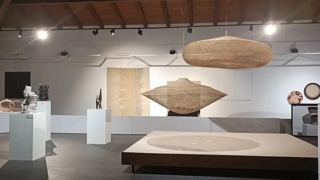 An interior view of an art gallery or exhibition space showcasing large, intricately woven sculptural pieces suspended from the ceiling and a textured art piece on the wall.