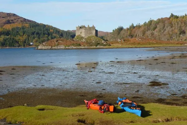 The image is well-lit and sharp, with good color accuracy, presenting a photo-realistic view of a castle by a loch. Key elements, including the castle and foreground kayaks, are in clear focus. The composition is generally strong, although the muddy foreshore and foreground kayaks slightly clutter the immediate view, preventing a perfect score.