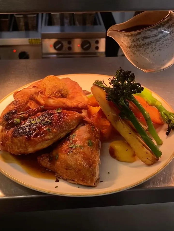 The image features a well-composed dish of roasted chicken and vegetables, enhanced by a dynamic pour of gravy. The main elements are sharp and well-lit, displaying accurate and appealing colors, contributing to a high degree of photo-realism. The background is a kitchen setting, which is slightly out of focus and does not detract significantly from the food.