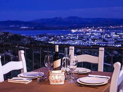An evening shot of a table set for dinner on a balcony or terrace, overlooking a city with twinkling lights, a body of water, and mountains in the background, all bathed in the blue hour light.