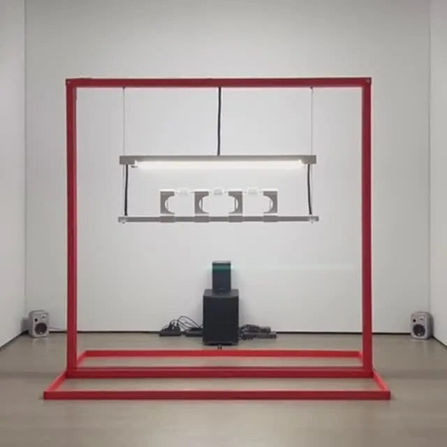 The image features a modern art installation, characterized by a striking red rectangular frame enclosing a setup of linear lighting fixtures, a speaker system, and other electronic components. The composition is clean and visually balanced, set against a minimalist white wall and concrete floor. The lighting is generally good, making the objects distinct, though the bright lights within the installation itself cause some minor glare. The image is sharp, photo-realistic, and exhibits good color accuracy, with all key elements in clear focus.