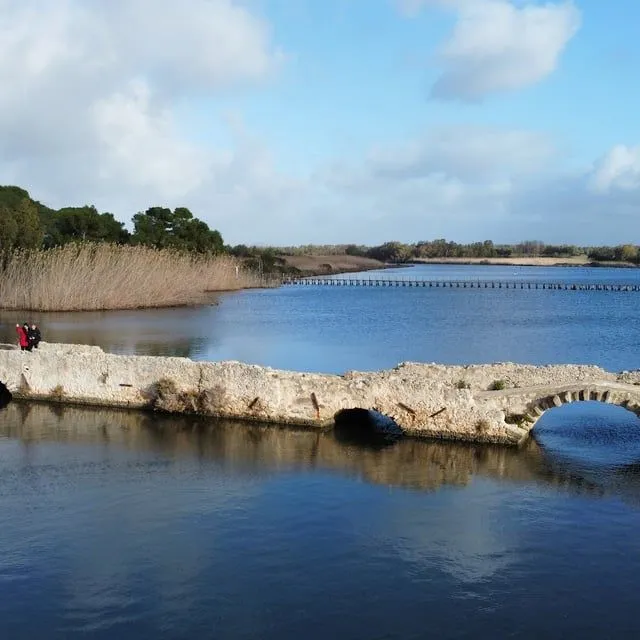 An aerial perspective of a tranquil water body traversed by an ancient-looking stone bridge. The water reflects the blue sky and scattered clouds, while one side of the bank is lined with tall reeds and trees. Two small figures are visible on the bridge.