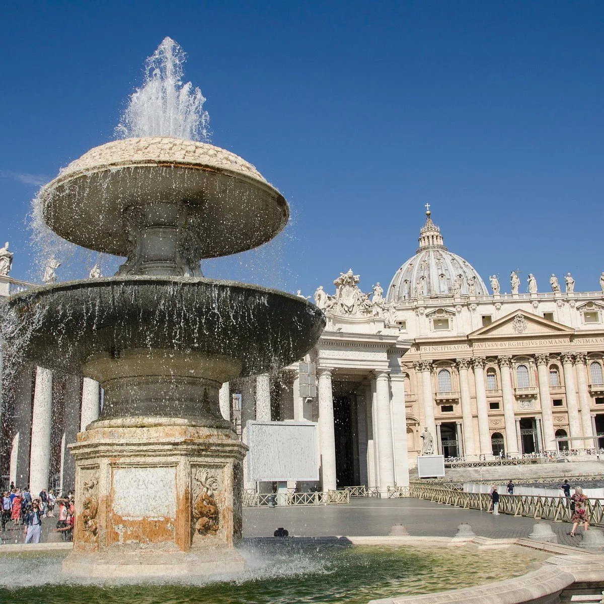 An impressive view of St. Peter's Square featuring a prominent fountain in the foreground and St. Peter's Basilica with its iconic dome in the background, under a clear blue sky. The fountain's water is actively flowing, and historical architecture dominates the scene.
