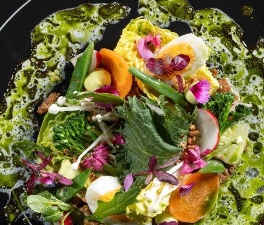 This image showcases a vibrant and artfully composed dish, excelling in its well-lit presentation and photo-realistic quality. The composition is strong, with key elements sharply in focus, highlighting the fresh and diverse ingredients like various greens, edible flowers, mushrooms, and boiled egg. Colors are accurate and rich, contributing to an appetizing visual. The swirling green sauce on the dark plate adds an artistic touch to the professional plating. No unwanted elements are present.