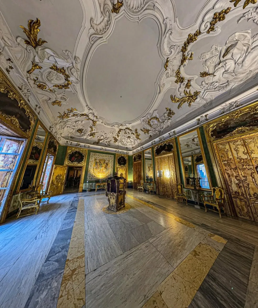 A grand and ornate interior space, featuring a highly decorated ceiling, gold-framed mirrors, and a polished marble floor, showcasing rich historical design.