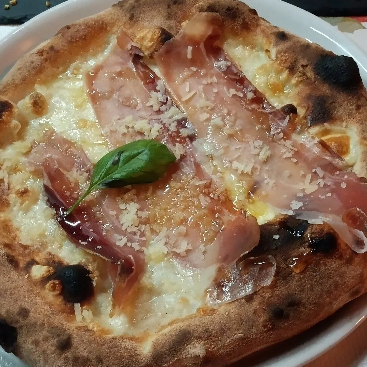 A close-up, top-down view of a freshly baked pizza topped with thinly sliced prosciutto, melted white cheese, a sprinkle of grated cheese, and a fresh basil leaf, presented on a white plate.