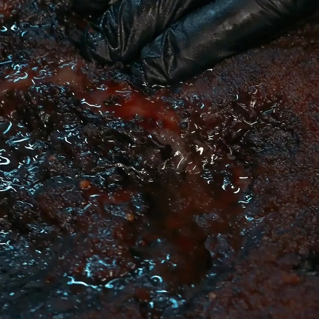 A very close-up shot of a dark, glistening piece of meat, likely smoked brisket, being pressed by a hand in a black glove, causing red juices to well up from the surface.