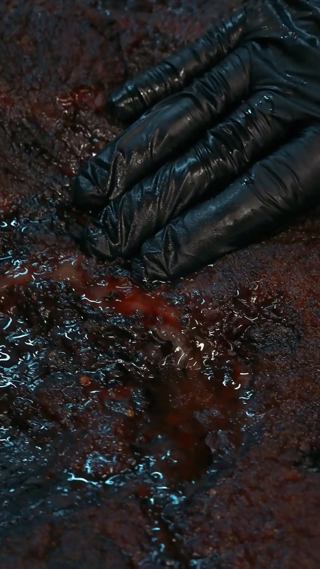 A very close-up shot of a dark, glistening piece of meat, likely smoked brisket, being pressed by a hand in a black glove, causing red juices to well up from the surface.
