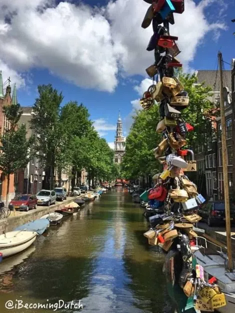 An image capturing a scenic view of an Amsterdam canal on a sunny day. The composition leads the eye down the canal towards a distant church steeple, with lush green trees, historic buildings, and boats lining the waterway. A prominent pole adorned with numerous love locks stands in the foreground, adding a distinctive element to the scene. The natural lighting is good, and the image is photo-realistic, though some areas could benefit from improved sharpness.