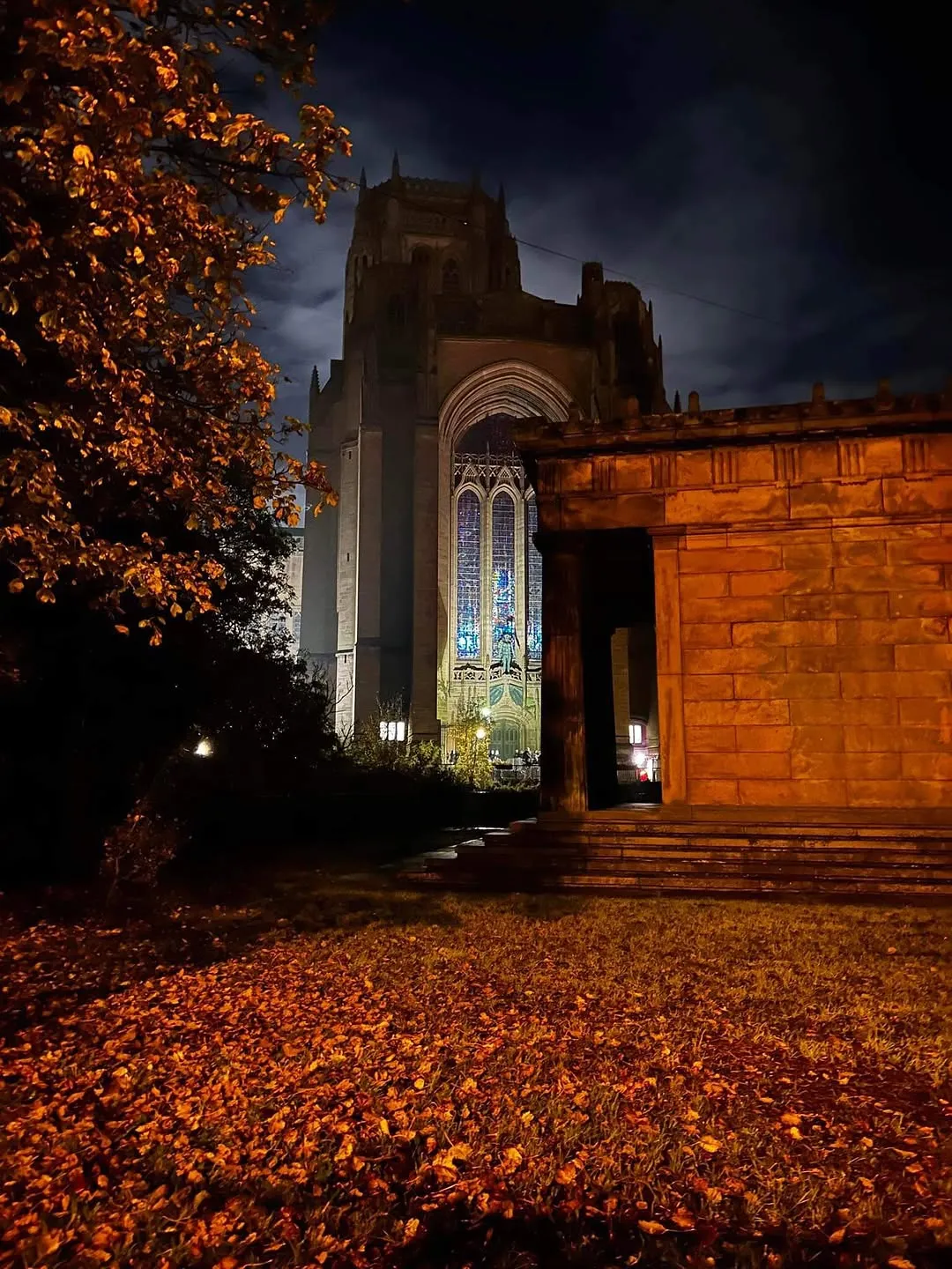 An atmospheric night shot of a cathedral with illuminated stained-glass windows, framed by autumn leaves and a stone structure.