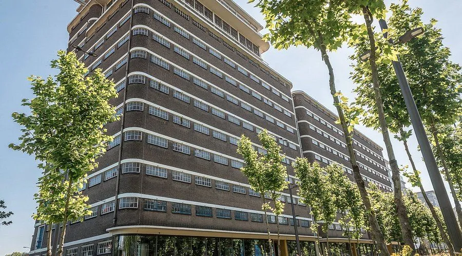 A well-composed and sharply focused photograph of a multi-story building with a distinctive curved architectural design. The image is well-lit by natural sunlight, showcasing the brickwork and numerous windows with accurate colors. Trees in the foreground provide natural framing.