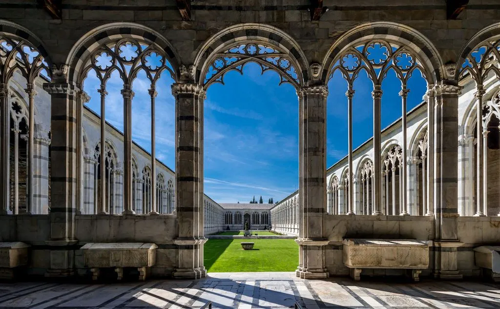 A striking architectural photograph capturing the interior view of a historic cloister or courtyard. Ornate Gothic arches in the foreground frame a meticulously maintained green lawn and the surrounding classical buildings under a clear, bright blue sky.