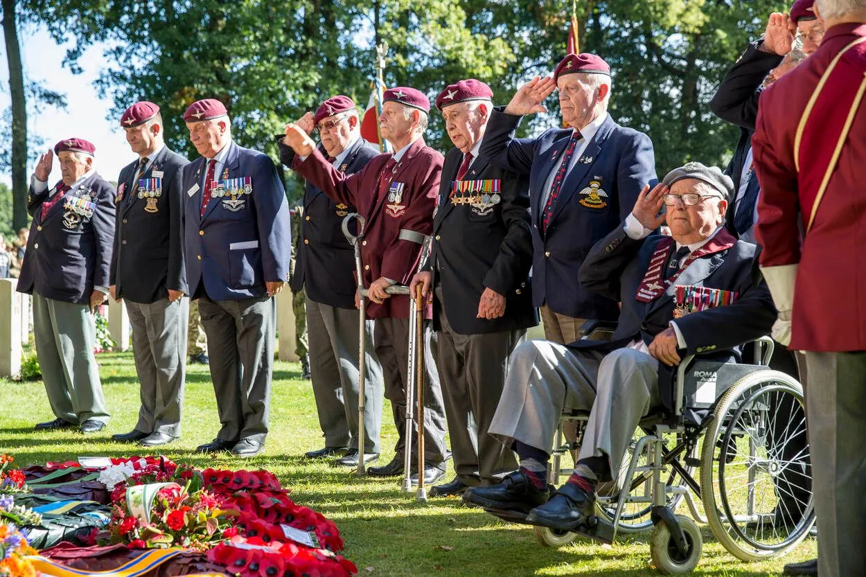 An outdoor scene depicting a group of elderly military veterans, many wearing berets and decorated uniforms, standing and sitting while saluting. Red poppy wreaths are visible in the foreground.