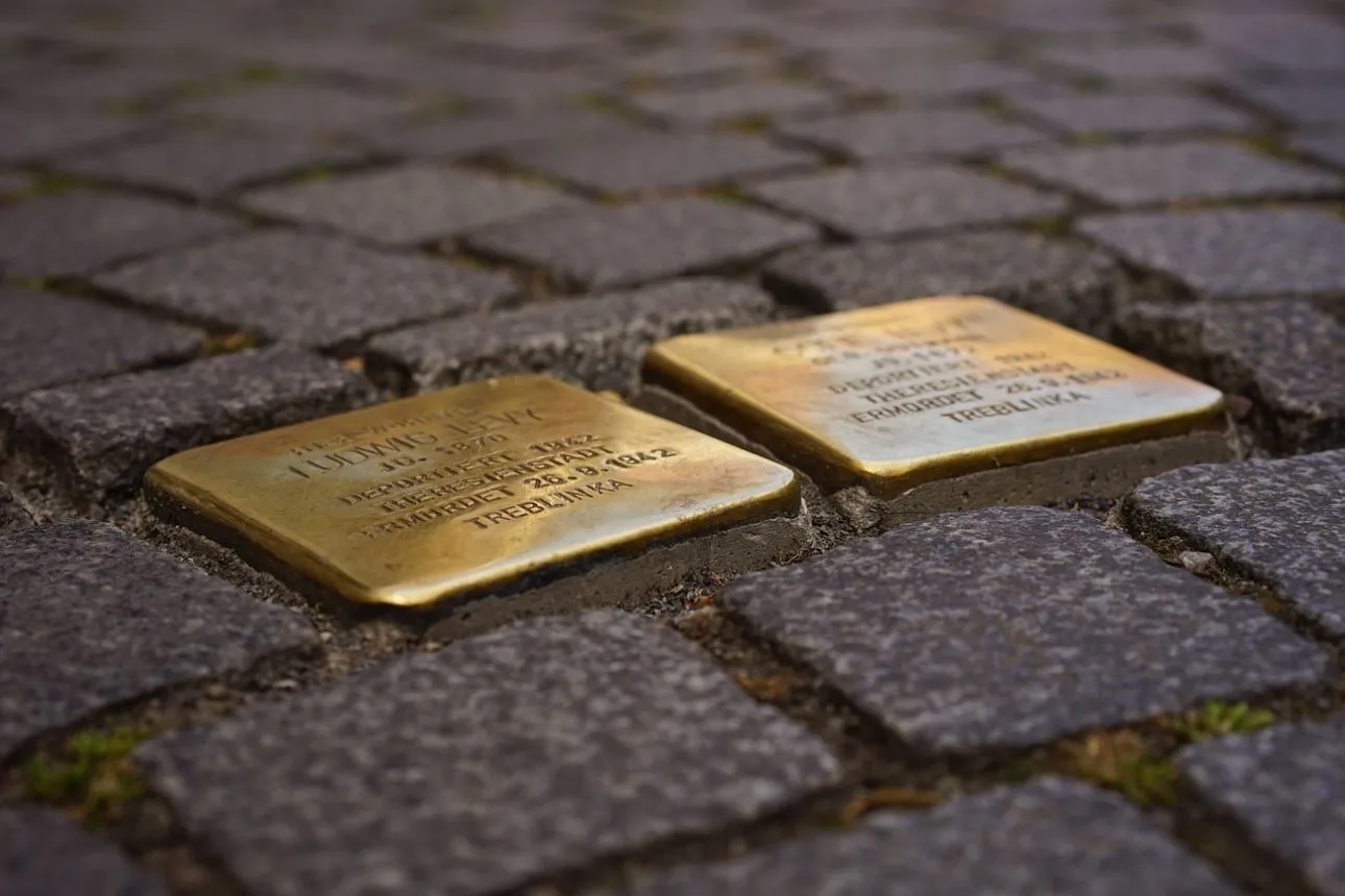 An editorial-style close-up photograph of two 'Stolpersteine' (stumbling blocks) embedded in a cobblestone street, with the nearest brass plaque clearly legible.