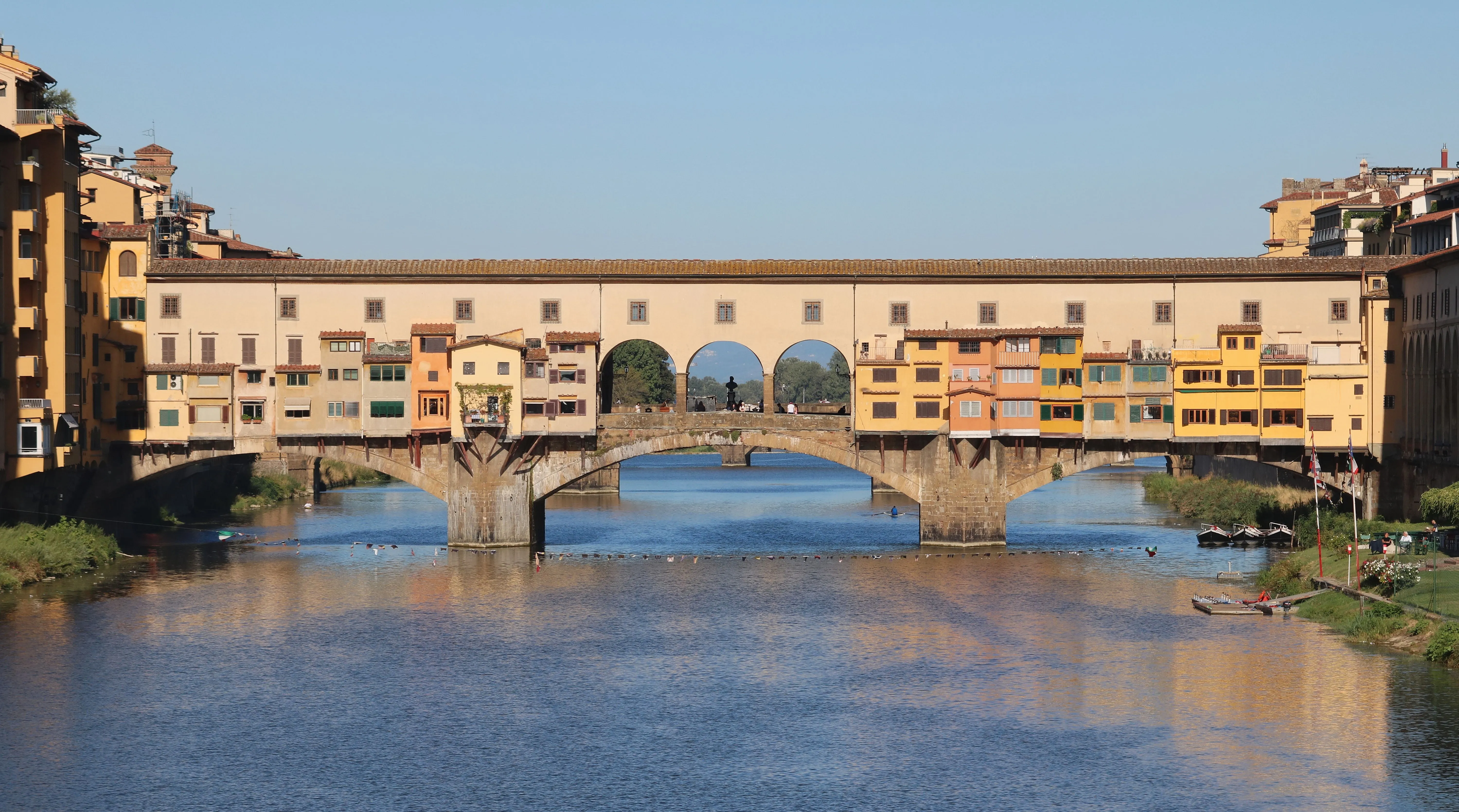 A beautiful and clear photograph of the historic Ponte Vecchio bridge spanning the Arno River in Florence, Italy, featuring the iconic shops built along its structure under a bright blue sky.