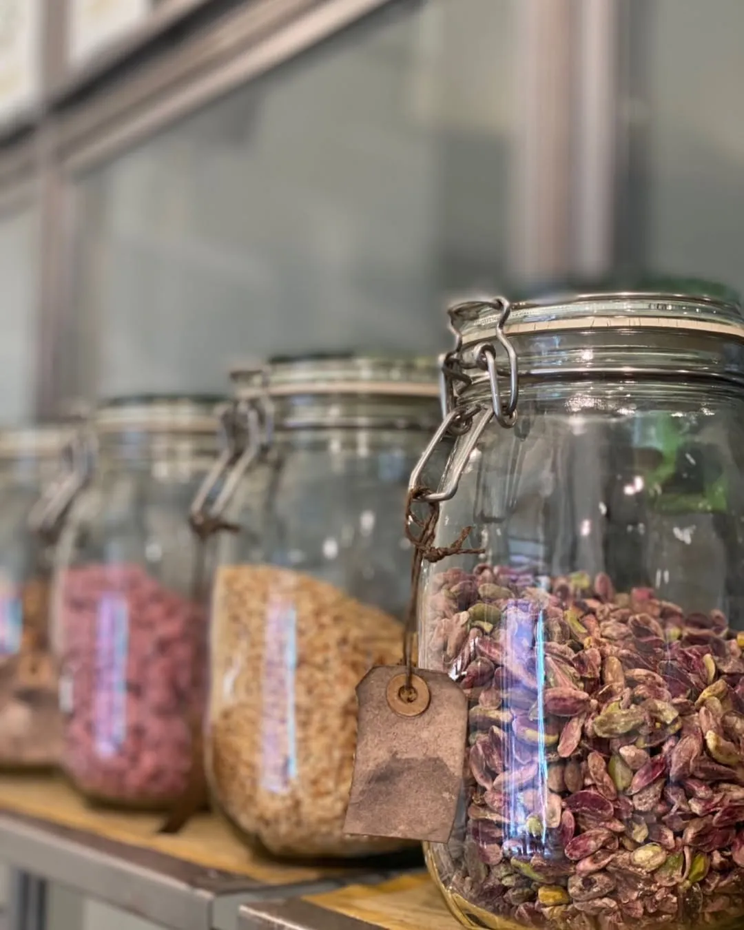 A close-up shot of several glass jars filled with various dry goods, including pistachios and other grains or seeds, neatly lined up on a shelf. The front jar is in sharp focus, while the background jars are softly blurred, creating depth.