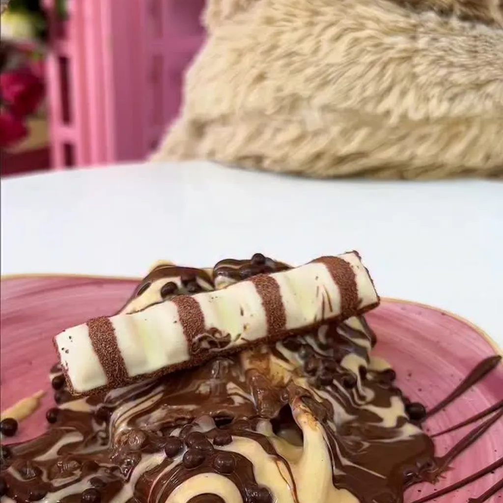 A close-up, high-quality image of a dessert featuring melted chocolate and cream, topped with a Kinder chocolate bar, served on a pink plate.