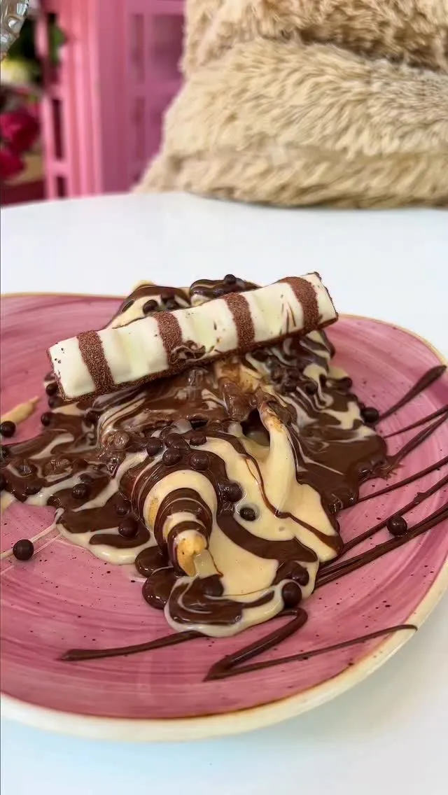 A close-up, high-quality image of a dessert featuring melted chocolate and cream, topped with a Kinder chocolate bar, served on a pink plate.