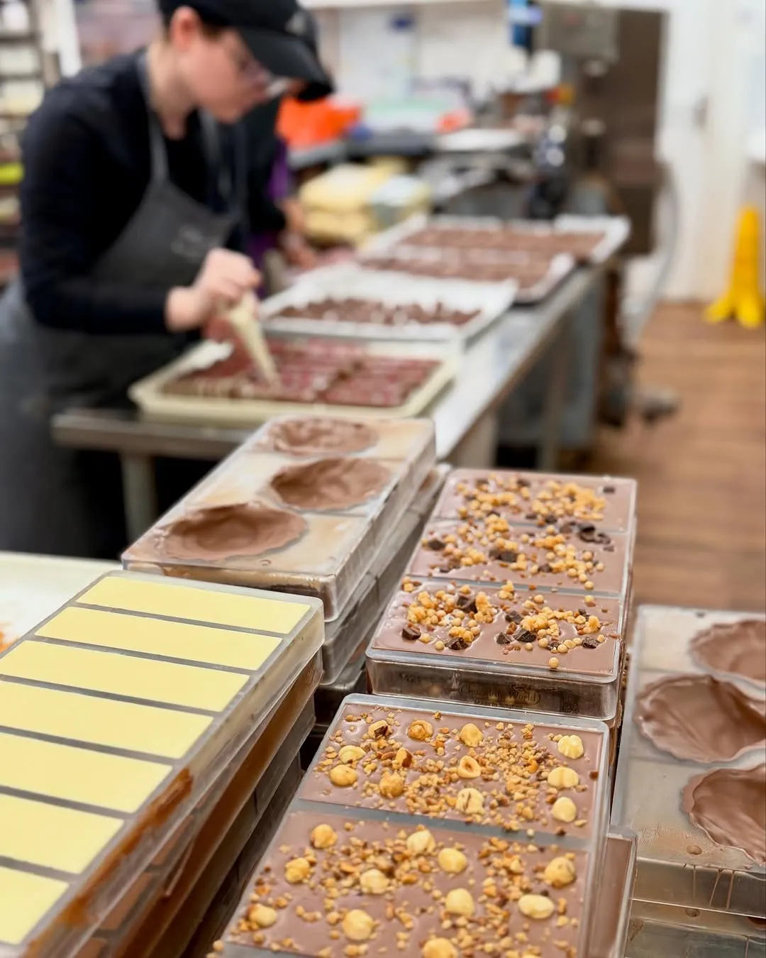 An image showcasing various chocolate bars and molds in the foreground, some with toppings like nuts, within a production or workshop setting. A person is visible in the blurred background, engaged in the chocolate-making process.