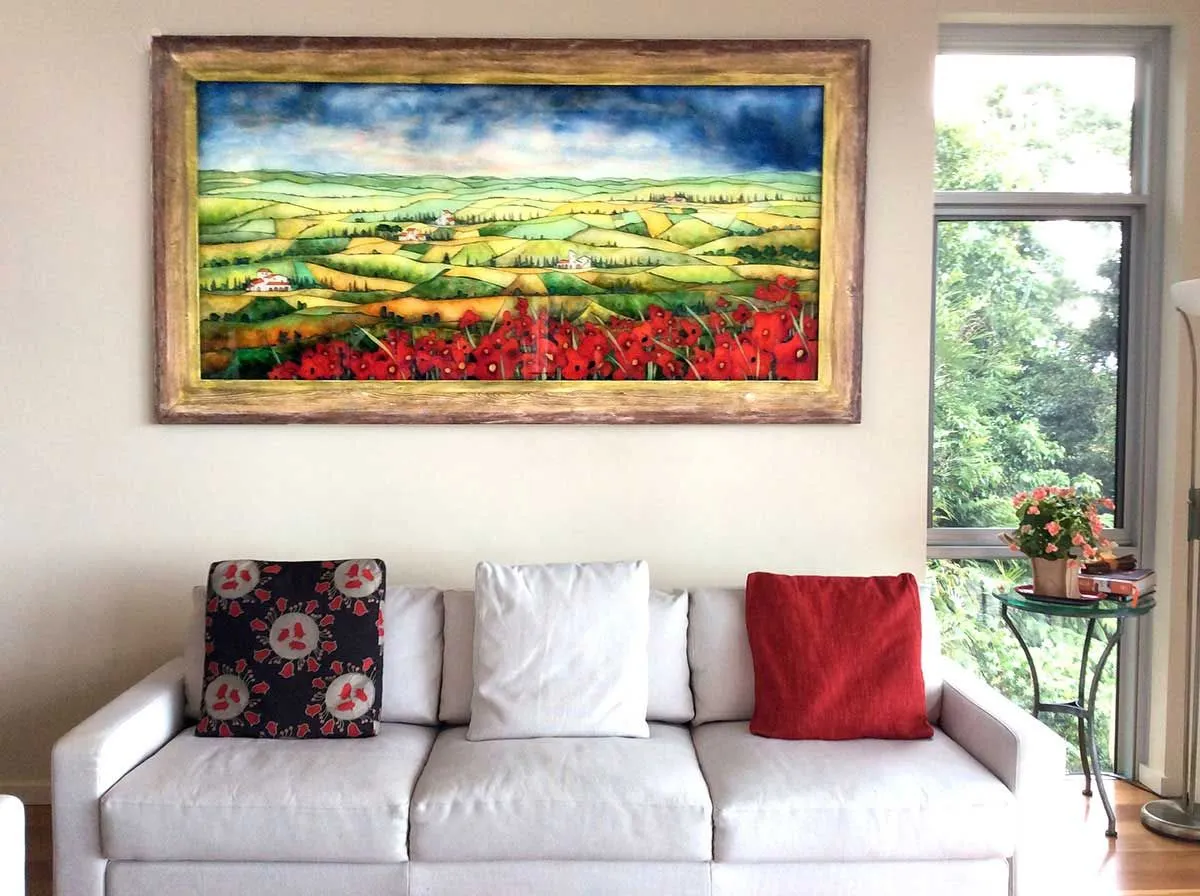 A large framed painting depicting a vibrant landscape with red poppies hangs prominently above a white sofa adorned with throw pillows in a modern living room setting, illuminated by natural light from a nearby window.