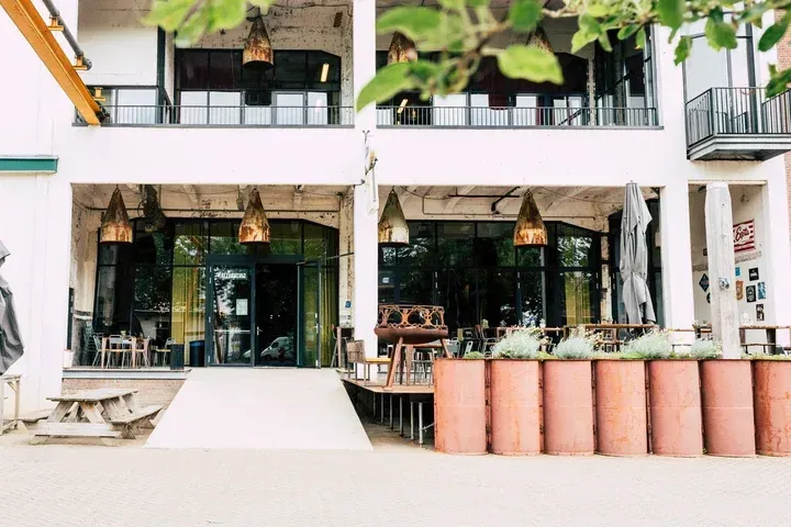 An editorial-style photograph capturing the modern, industrial exterior of a building, likely a restaurant or cafe, featuring an outdoor seating area. The composition is balanced with foreground foliage, a central building, and unique rusted barrel planters. The lighting is soft and even, and the colors are natural and true to life.