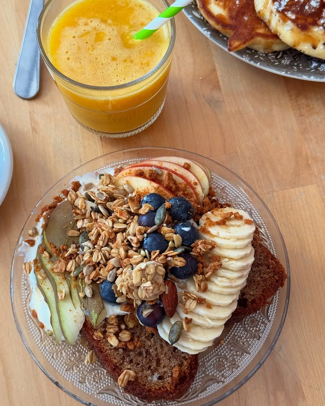 A well-lit and sharp image of a healthy breakfast, featuring bread topped with granola, fresh fruits (banana, apple, blueberry, pear), and a glass of orange juice. The colors are natural and vibrant, contributing to a photo-realistic appearance. The composition is generally good, though a minor part of another plate is visible in the background.