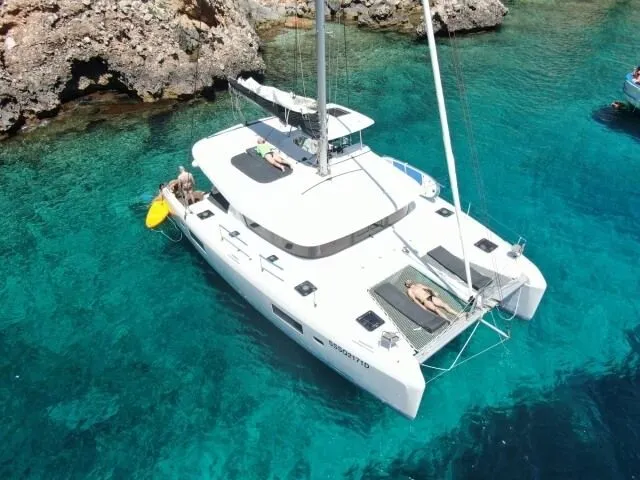 A high-angle view of a white catamaran anchored in crystal-clear turquoise waters next to a rocky coast, with people relaxing on board.