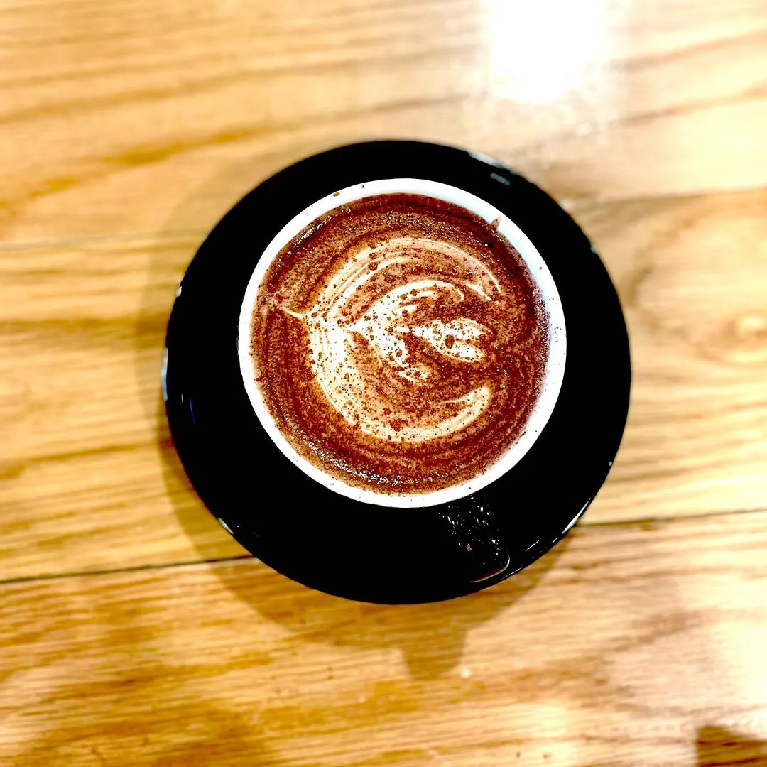 A well-composed, top-down shot of a hot beverage, likely coffee or hot chocolate, with intricate latte art on a black saucer. The drink is centered on a light wooden table, which provides a clean and natural background.