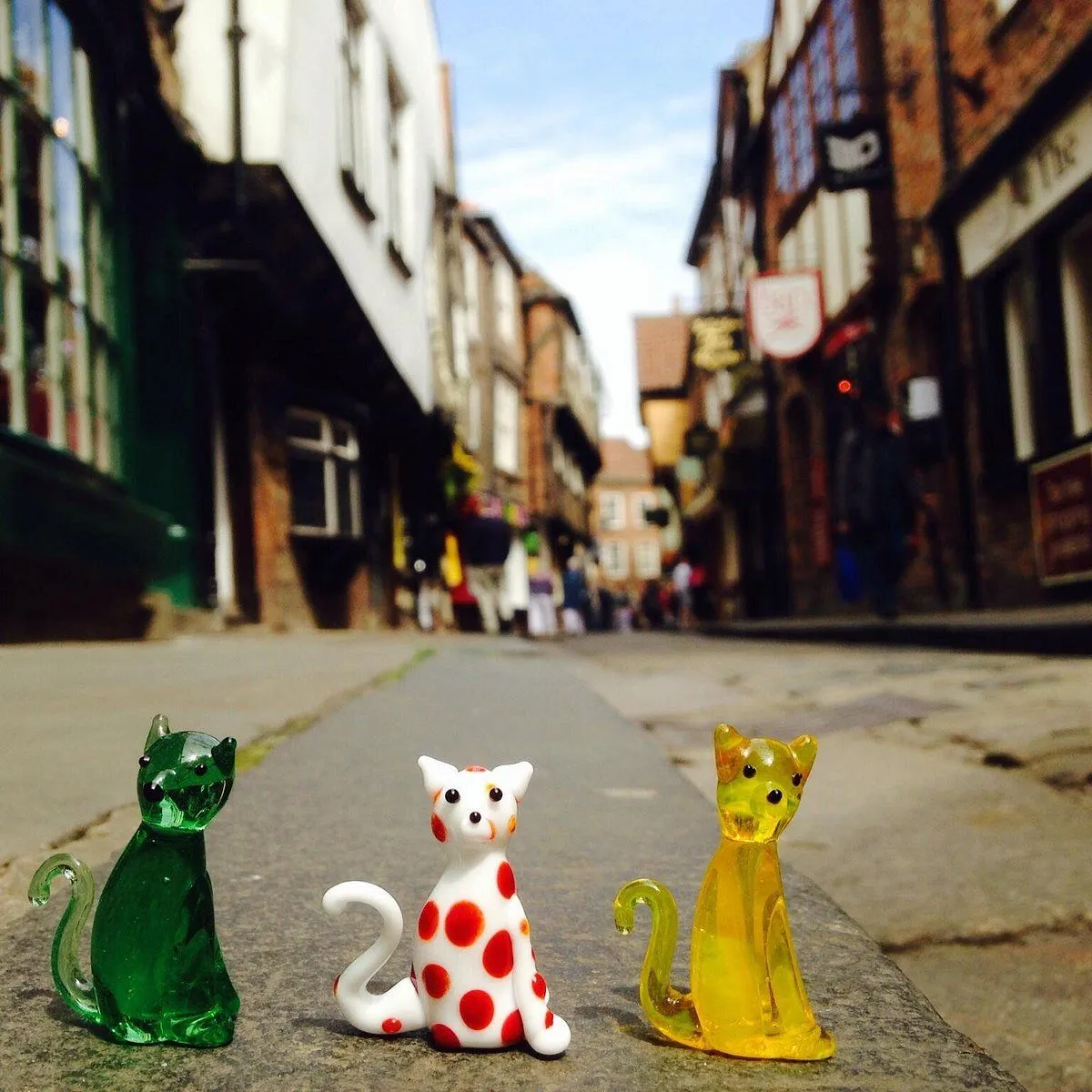 The image features three colorful glass cat figurines in sharp focus, set against a charming, blurred street background. The composition effectively uses depth of field to highlight the main subjects. Lighting is natural and colors are vibrant and accurate.