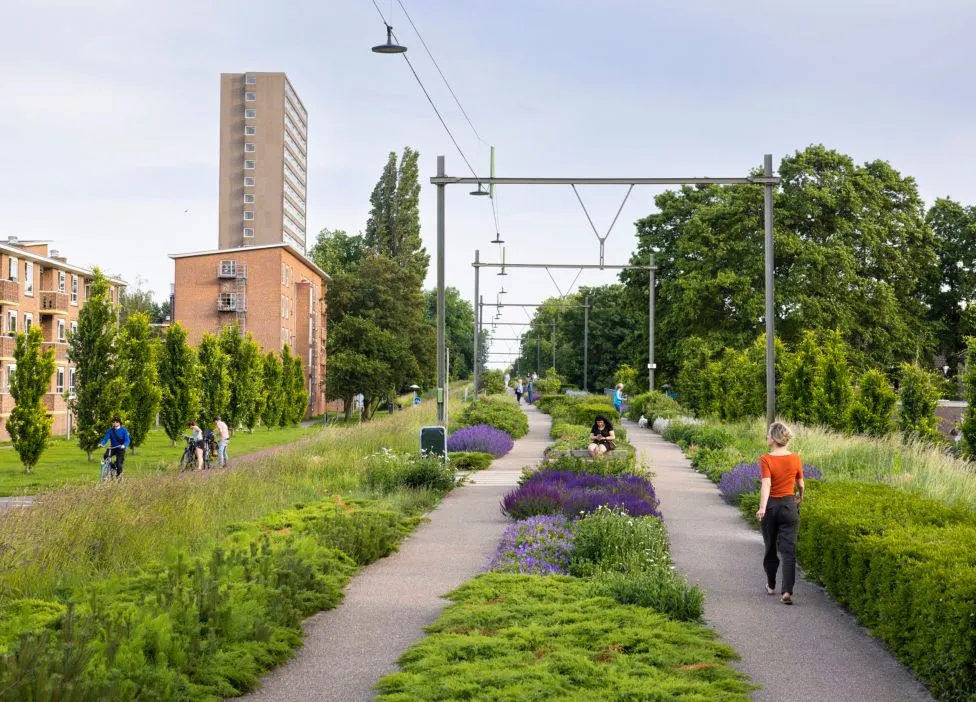 An elevated perspective of an urban green space featuring winding pedestrian paths flanked by lush green shrubs and vibrant purple flowers. Overhead structures, reminiscent of a former railway or tram line, span the path, leading the eye into the distance where people are walking and cycling. A tall residential building and a brick structure are visible in the background amidst abundant greenery.