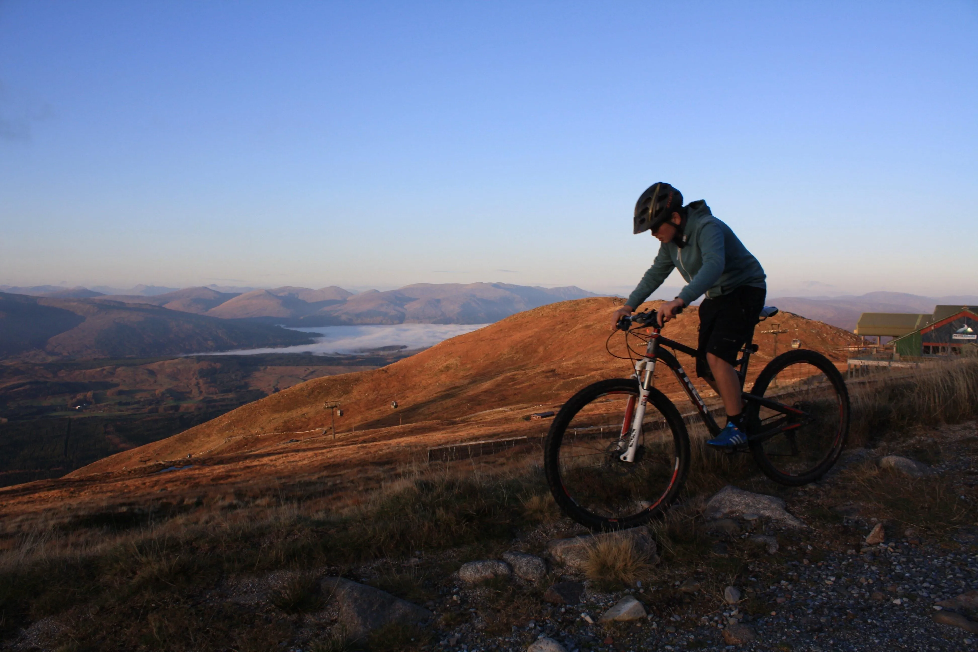 A person on a mountain bike is captured on a golden-hued hillside, overlooking a misty valley and distant mountains under a clear sky. The scene is set during what appears to be either sunrise or sunset, casting warm light across the landscape.