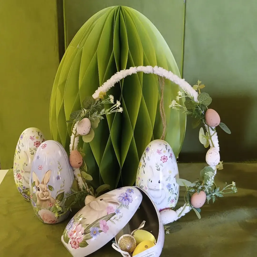 The image features a collection of Easter decorations, including several intricately painted eggs with bunny and floral designs, a decorative wreath, and a large green honeycomb paper egg as a backdrop. The items are well-arranged, creating an appealing and festive display.