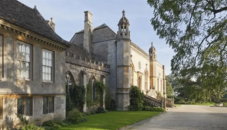 The image features a stately, historic building with ornate architectural details, likely a manor or castle wing. The natural sunlight illuminates the stone facade, creating good contrast and highlighting the intricate design elements. The composition frames the building effectively, with a well-maintained lawn and pathway in the foreground. The image is sharp, color-accurate, and highly photo-realistic, with all key elements in clear focus.