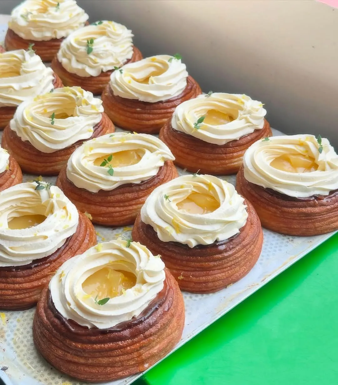 A well-composed and brightly lit close-up image of multiple pastries arranged neatly on a tray. The pastries feature a layered, golden-brown base topped with piped white cream, a yellow fruit filling in the center, and garnished with small green leaves. The overall sharpness and color accuracy contribute to a highly appealing and photo-realistic presentation.