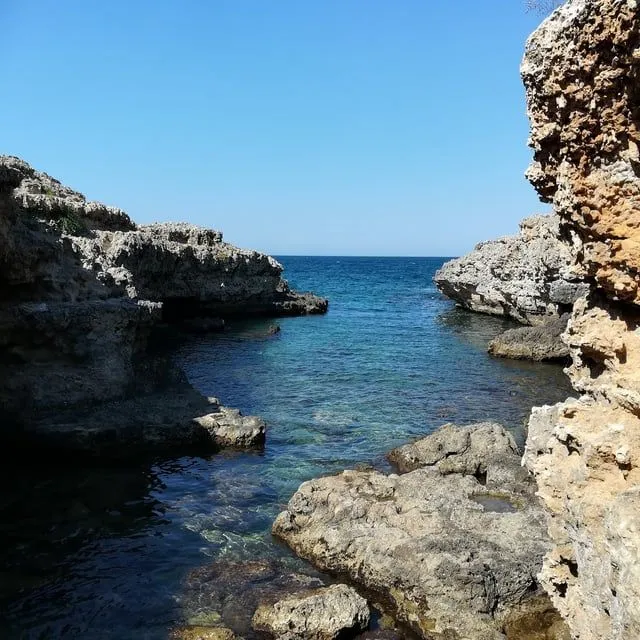 An aerial view of a person standing on a rocky outcrop overlooking a clear turquoise sea.