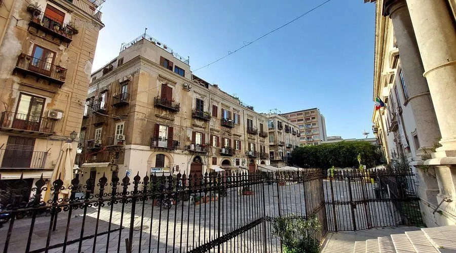 An editorial style photograph capturing an urban scene featuring aged buildings with balconies and shutters under a clear blue sky. A decorative metal fence dominates the foreground, partially obscuring the view of the street and the architecture beyond. The scene is well-lit by daylight.