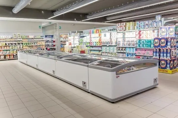 An interior view of a bright and organized supermarket aisle featuring a long line of white freezer display cases on the left and densely stocked shelves with various products on the right, under modern overhead lighting.
