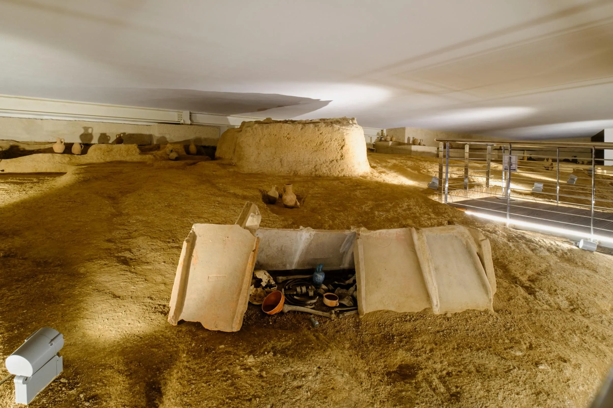 An image of an indoor archaeological excavation site, showcasing an open burial or structural remains with artifacts, surrounded by unearthed earth and other excavated features.