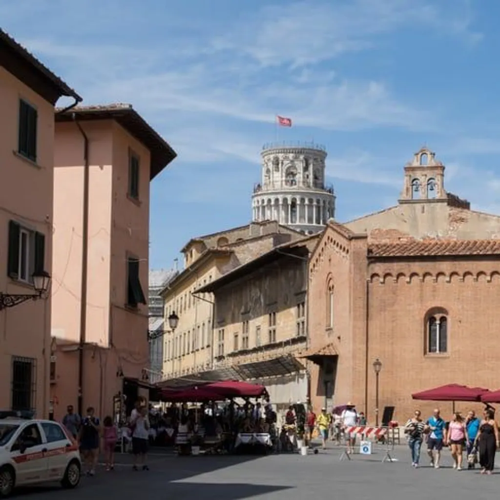 An editorial-style image capturing a street scene with historic architecture, including a prominent tower in the background, market stalls, and people.