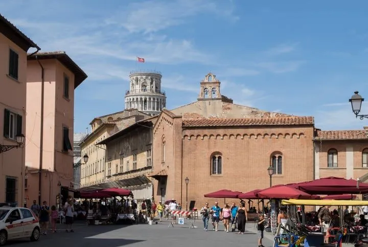 An editorial-style image capturing a street scene with historic architecture, including a prominent tower in the background, market stalls, and people.