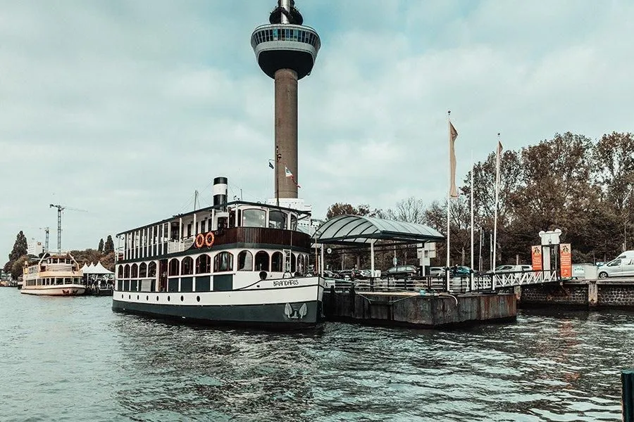 An image featuring a historic-looking boat docked near a modern observation tower, with water in the foreground and a cloudy sky. The scene depicts an urban waterfront.