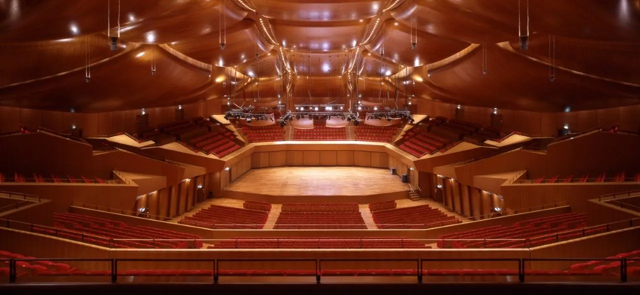 The image features a well-composed, wide shot of an empty concert hall or auditorium. The composition is strong, showcasing the stage, the tiers of red seating, and the unique wooden ceiling architecture. Lighting is excellent, illuminating the entire space evenly and highlighting the textures of the wood and fabric. Colors appear accurate and natural. The image is photo-realistic, and key elements such as the stage and seating areas are sharp and in focus. There are no unwanted elements present.