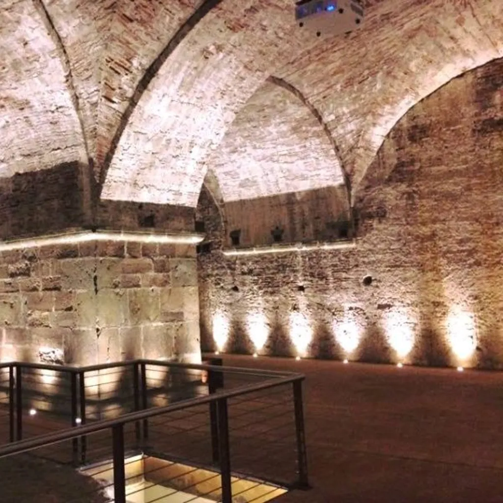 An interior shot of an ancient or historic vaulted space, likely a cellar or crypt, constructed from stone and brick. The space is dramatically illuminated by spotlights on the floor and linear lights along the pillars, highlighting the architectural details. A metal railing with wire infill provides a foreground element, suggesting a pathway or viewing area. A projector is visible hanging from the ceiling in the upper left.