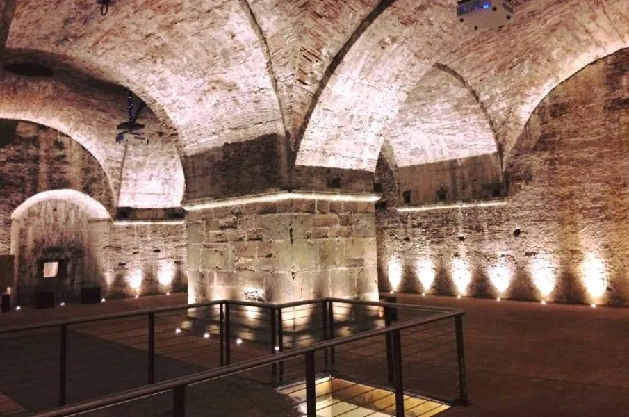 An interior shot of an ancient or historic vaulted space, likely a cellar or crypt, constructed from stone and brick. The space is dramatically illuminated by spotlights on the floor and linear lights along the pillars, highlighting the architectural details. A metal railing with wire infill provides a foreground element, suggesting a pathway or viewing area. A projector is visible hanging from the ceiling in the upper left.